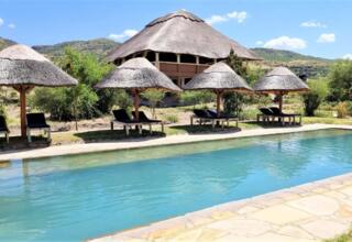 Outdoor pool with stone deck surrounded by thatched umbrellas and lounge chairs, set against green hills under a clear blue sky