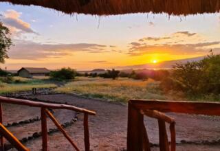 Sunset view from a rustic lodge porch overlooking grassy plains, distant thatched cabins, and a tranquil horizon