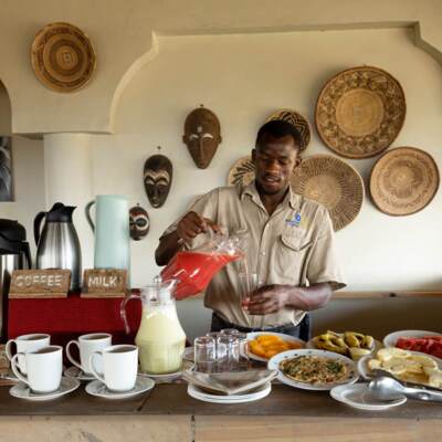 Hotel staff pours fresh juice at breakfast buffet with coffee, milk, fruit, pancakes, and bread in a rustic setting with African decor.