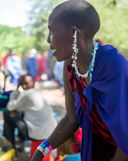 Woman wearing traditional Maasai clothing and jewelry, engaged in outdoor cultural gathering with people in colorful attire.