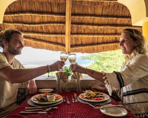 Couple toasting with wine glasses at an open-air thatched roof dining area with a scenic nature view