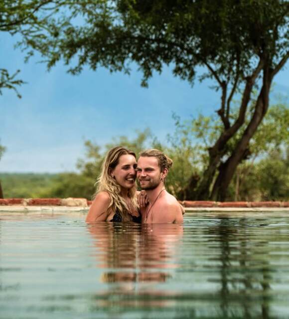 Couple enjoying a serene swim in an outdoor pool surrounded by trees and natural greenery under a clear sky