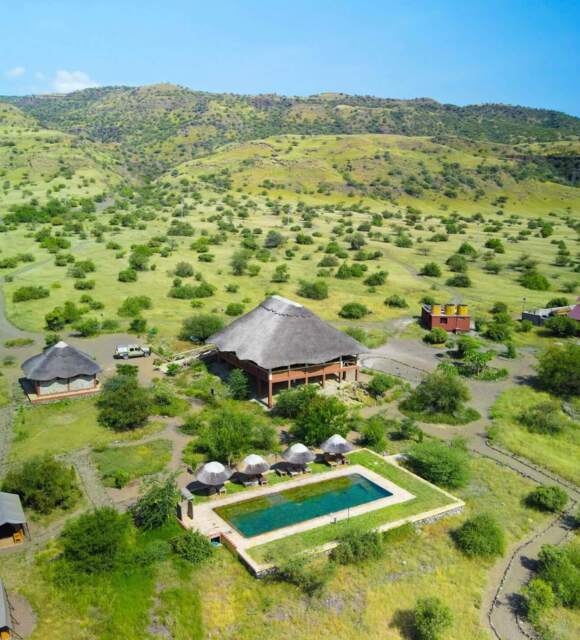 Eco-lodge with thatched roofs and a rectangular pool surrounded by lounge chairs, set in rolling green hills under a clear blue sky