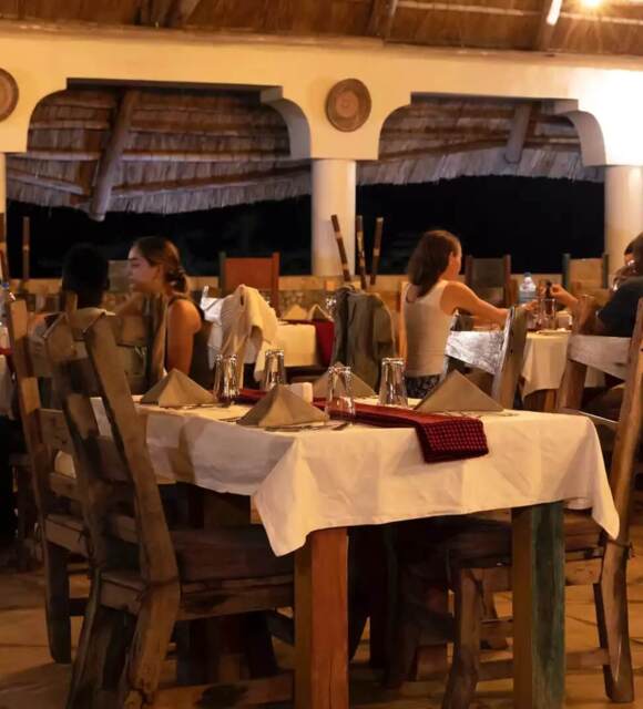 Open-air restaurant with thatched roof, wooden chairs, stone floor, and guests dining in warm, intimate evening ambiance