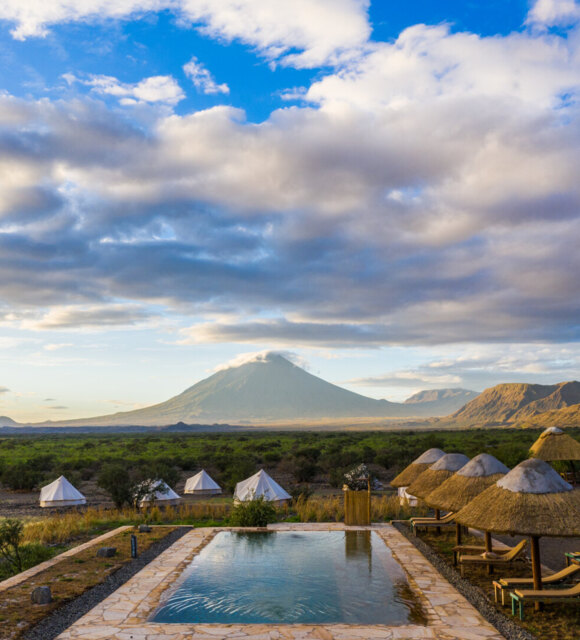 Outdoor pool with stone deck and thatched umbrellas beside lounge chairs, overlooking tents, lush plains, and a distant volcanic mountain