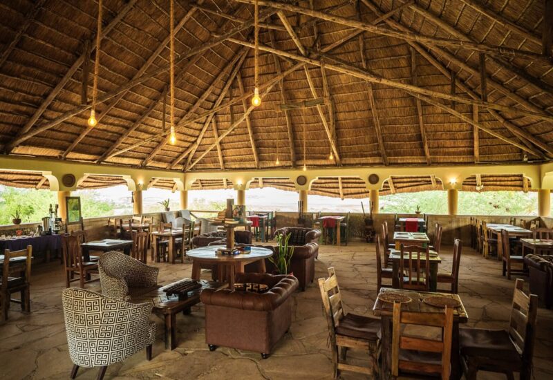 Open-air safari lodge dining area with thatched roof, wooden tables, leather armchairs, and views of lush greenery outside