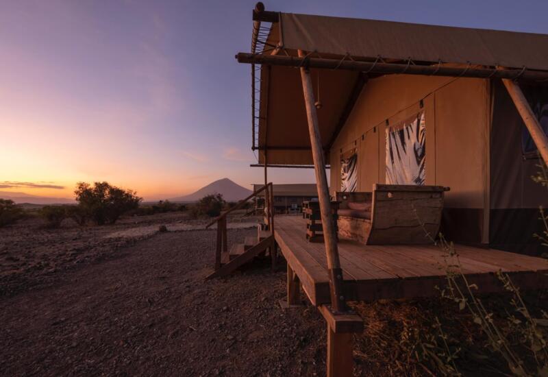 Rustic safari tent with wooden deck seating at sunset, overlooking a vast savannah with distant mountain views and soft evening light