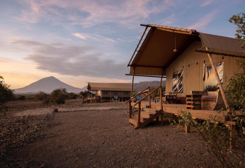 Luxury safari tents with wooden decks and seating areas set in a rugged landscape, with a mountain peak under a colorful sunset sky