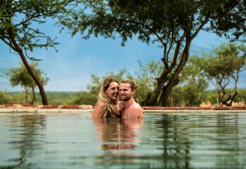 Couple enjoying a serene swim in an outdoor pool surrounded by trees and natural greenery under a clear sky