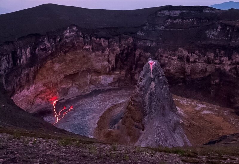 View of an active volcanic crater at dusk with glowing lava streams and rugged rocky walls surrounding the volcanic cone