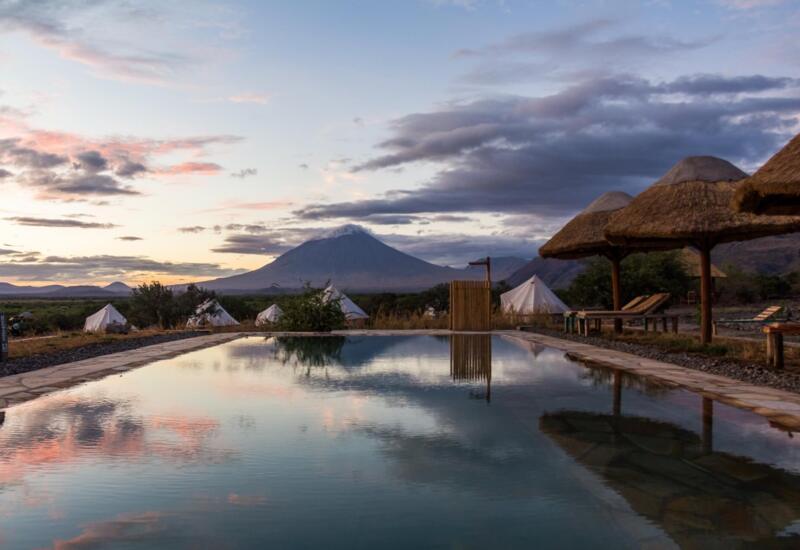 Serene infinity pool at a safari lodge with thatched umbrellas, lounge chairs, and white tents against a mountain sunset backdrop