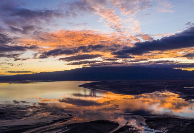 Serene lakeside view at sunset with dramatic clouds reflecting vibrant orange and purple hues over calm water and distant mountains