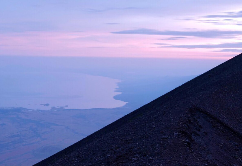 Twilight view from a mountain peak overlooking a calm body of water and distant shore under a purple sky