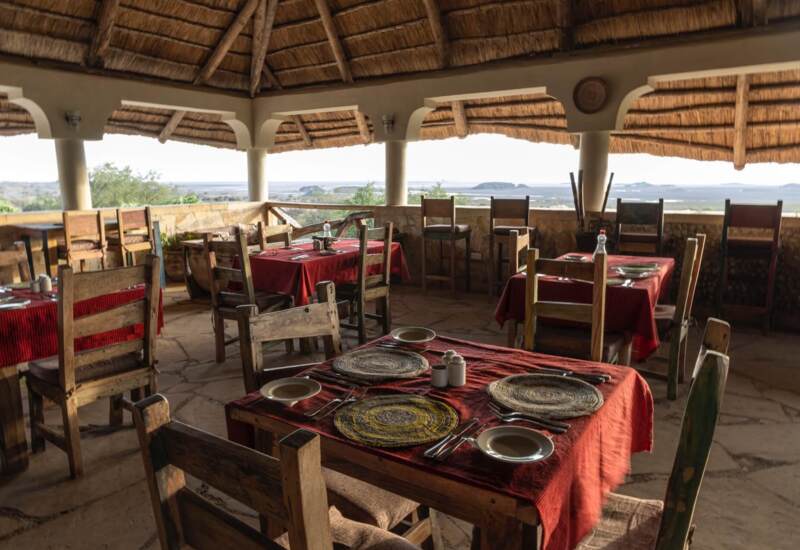 Open-air restaurant with thatched roof, rustic wooden tables with red tablecloths, woven placemats, and scenic nature views