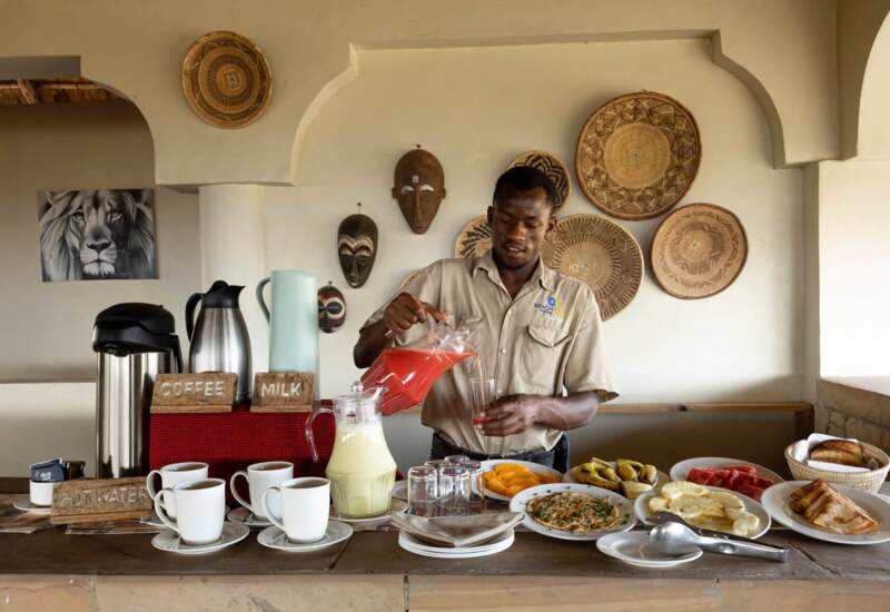 Hotel staff pours fresh juice at breakfast buffet with coffee, milk, fruit, pancakes, and bread in a rustic setting with African decor.