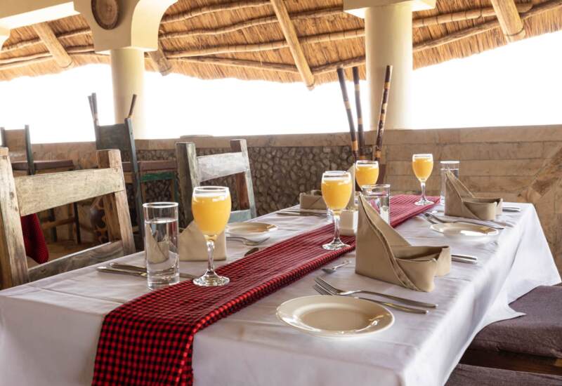 Rustic open-air dining area with wooden chairs, white tablecloth, red checkered runner, and glasses of orange juice set for guests