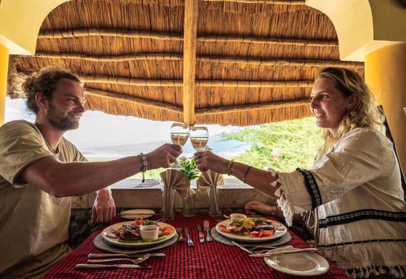 Couple toasting with wine glasses at an open-air thatched roof dining area with a scenic nature view
