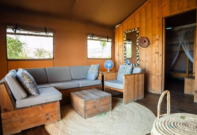 Cozy lounge area with wooden furniture, gray cushions, patterned pillows, and a rustic coffee table on a woven rug in a tent-style room.