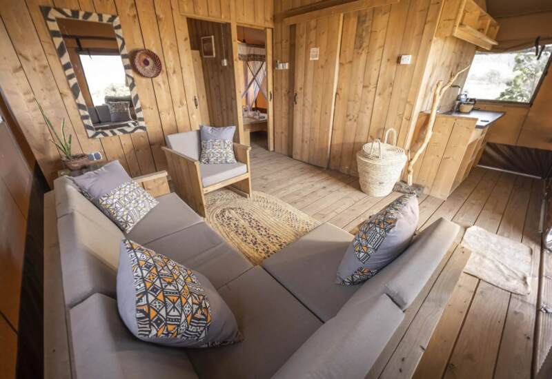 Cozy wooden lounge area with soft grey cushions, patterned pillows, woven rug, natural decor, and adjoining bedroom with canopy bed