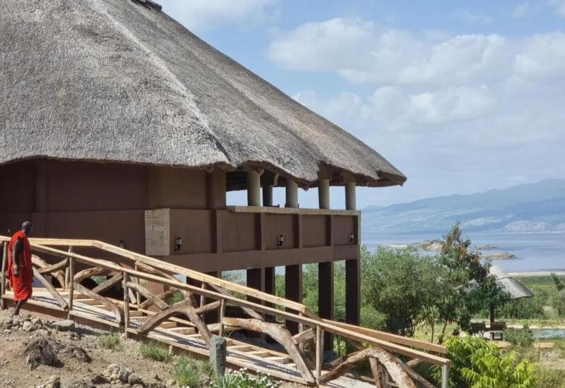 Thatched roof lodge with elevated wooden walkway overlooking a serene lake and distant mountain range under a partly cloudy sky