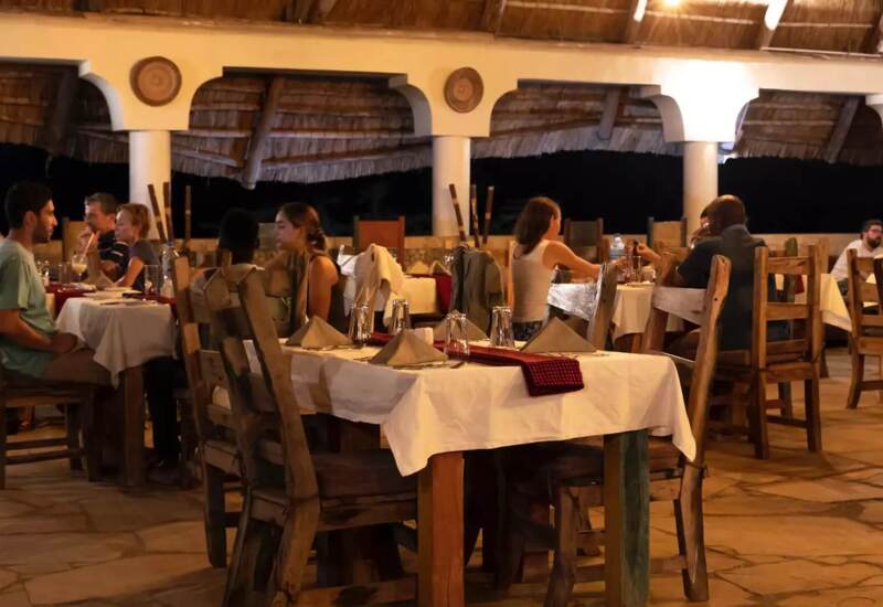 Open-air restaurant with thatched roof, wooden chairs, stone floor, and guests dining in warm, intimate evening ambiance