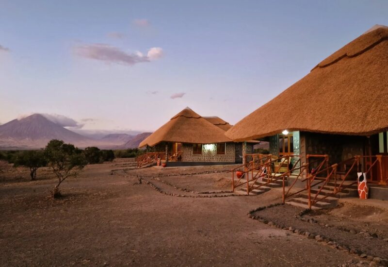 Lodge with thatched roofs and stone walls set in a dry landscape, featuring wooden railings and views of a distant volcanic mountain