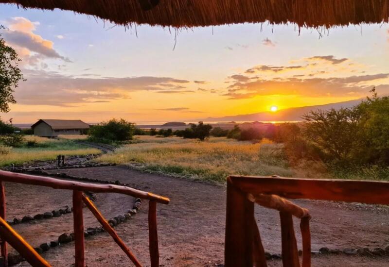 Sunset view from a rustic lodge porch overlooking grassy plains, distant thatched cabins, and a tranquil horizon