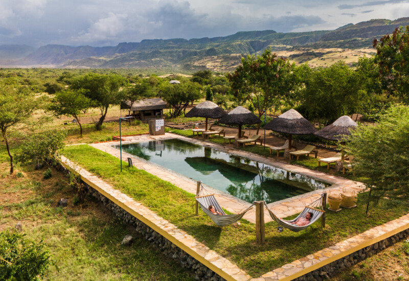 Secluded outdoor pool with stone deck, thatched umbrellas, lounge chairs, and two guests relaxing in hammocks amid scenic mountain landscape