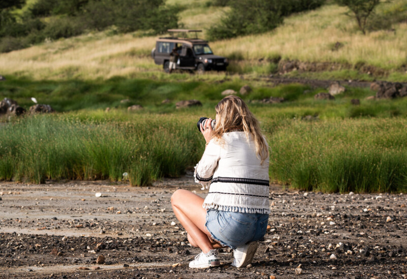 Guest in casual outfit photographing nature near safari vehicle in a grassy, rocky outdoor landscape