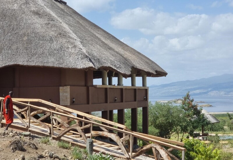 Thatched roof lodge with rustic wooden railings overlooking a serene lake and distant mountains under a partly cloudy sky