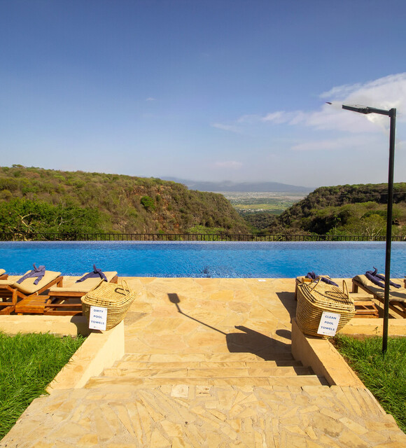 Infinity pool on a stone terrace with wooden loungers, umbrellas and towel baskets, overlooking a green valley and distant plains.