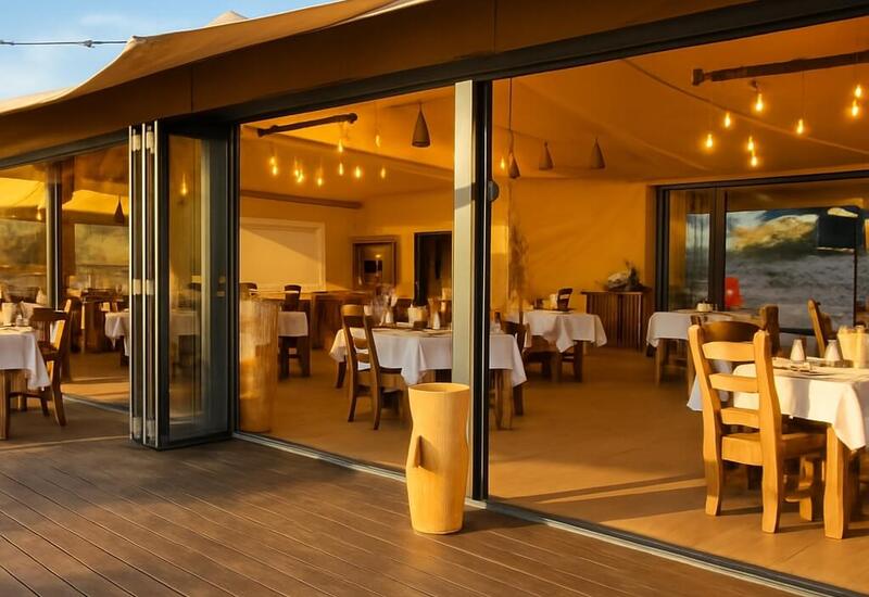 Open-air beachfront restaurant with sliding glass doors, wooden chairs, white linens, warm string lights and ocean views.