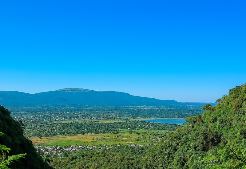 Panoramic hilltop view over green valley and lakeside village toward a flat-topped mountain under a clear blue sky