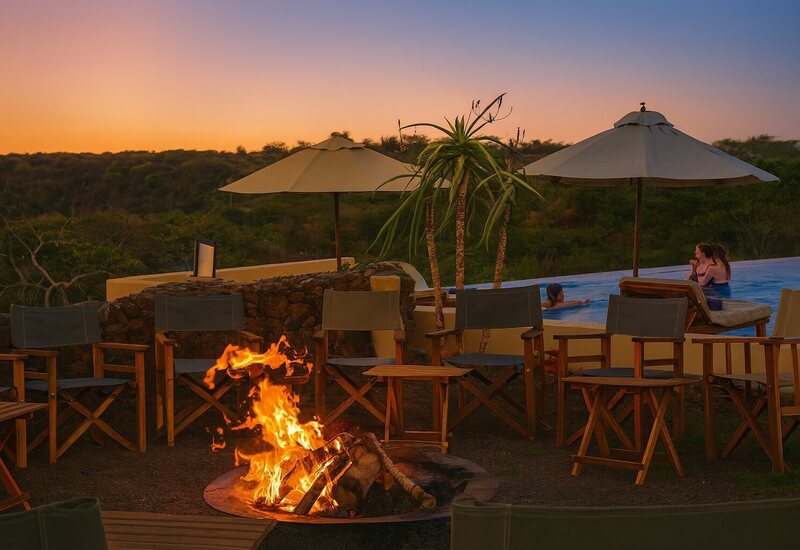 Sunset poolside with infinity pool and umbrellas, guests in water beside wooden loungers and a glowing fire pit.