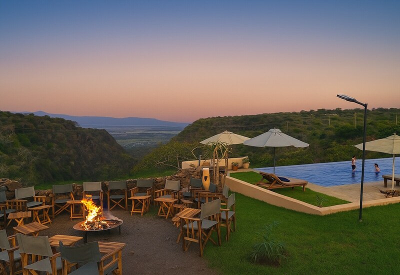 Sunset terrace with lit fire pit surrounded by chairs, infinity pool with two guests wading, sunbeds and umbrellas over a green valley.
