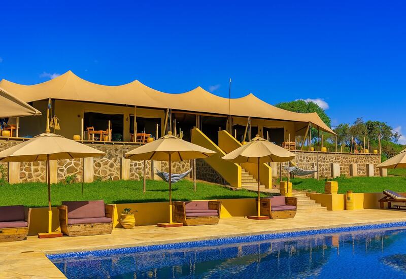 Tented luxury lodge with shaded veranda, stone terrace and hammocks above a pool with umbrellas, wooden lounge seats and blue sky.