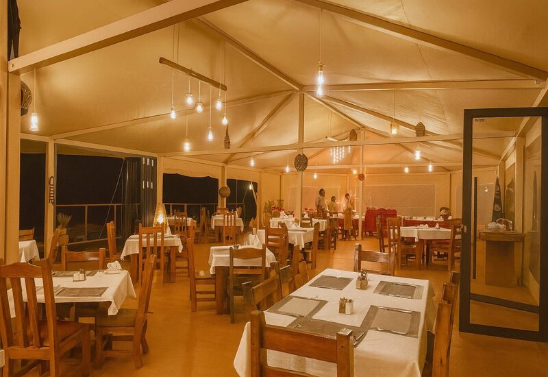 Tented restaurant with wooden tables, white linens, warm hanging bulbs and staff preparing a buffet.