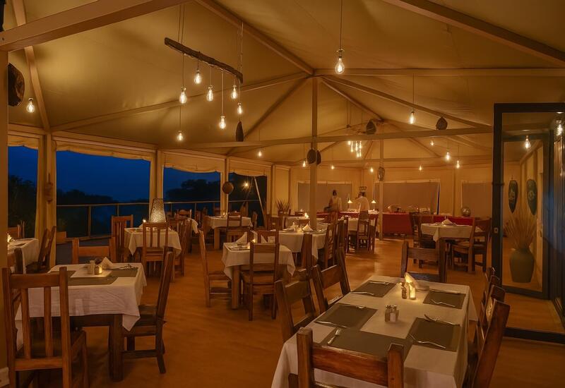 Warm lodge dining under a canvas tent with wooden tables, candlelit settings, hanging Edison bulbs and a buffet station overlooking dusk