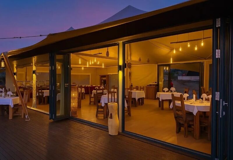 Tented open-air restaurant at dusk with warm hanging bulbs, wooden tables dressed in white linens on an open wooden deck.
