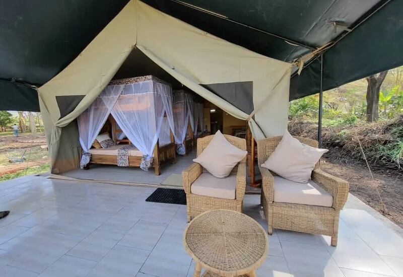Safari-style canvas tent with wooden four-poster beds draped in mosquito nets, shaded tiled terrace with wicker armchairs and small table.