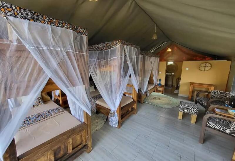 Spacious safari-style tent with three wooden canopy beds draped in white mosquito nets, patterned bedding, lounge chairs and rustic sink area.