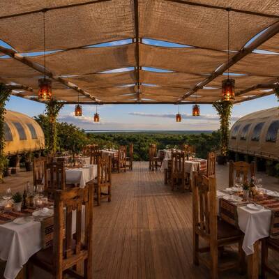 Open-air treetop dining deck with rustic wooden tables, white linens, hanging lanterns and vine-wrapped columns overlooking forest at sunset.