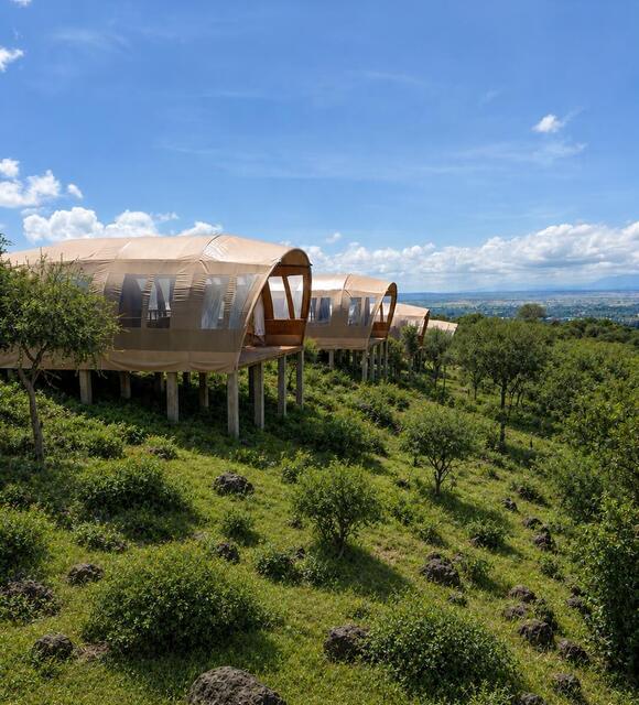 Elevated luxury glamping tents on stilts with wooden decks overlooking a green hillside and panoramic valley under a bright blue sky