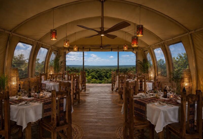 Tented fine-dining room with rustic wooden tables, lantern lighting and open front overlooking lush treetops and distant horizon.