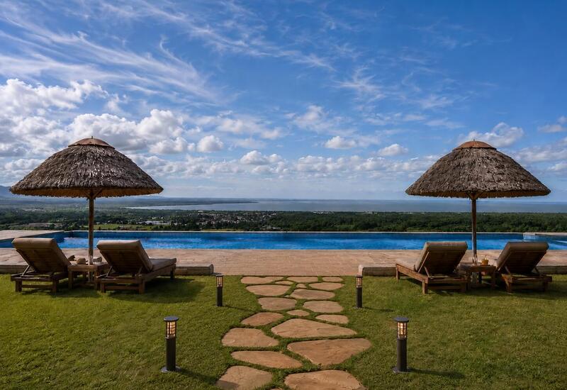 Infinity pool with panoramic lake view, two wooden loungers under thatched parasols, stone path and lawn-lit terrace.