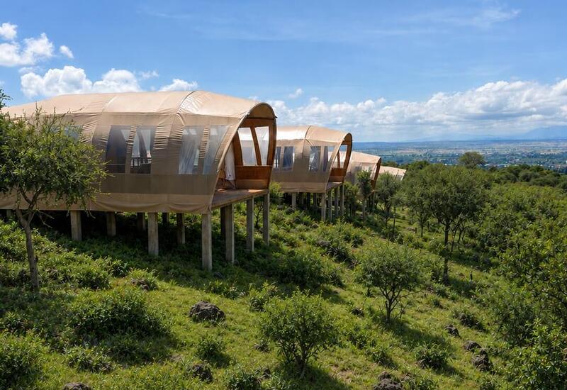 Elevated luxury glamping tents on stilts with wooden decks overlooking a green hillside and panoramic valley under a bright blue sky