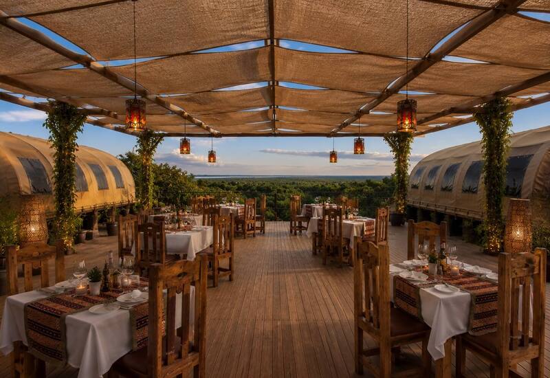 Open-air treetop dining deck with rustic wooden tables, white linens, hanging lanterns and vine-wrapped columns overlooking forest at sunset.