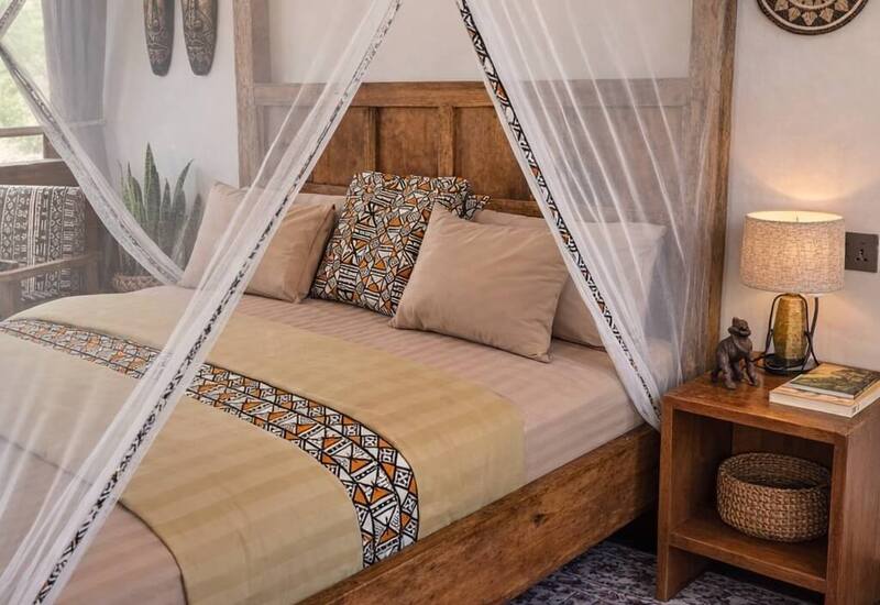 Wooden four‑poster canopy bed with sheer mosquito net, patterned pillows and throw, bedside lamp and woven decor in a cozy boho-style guest room.