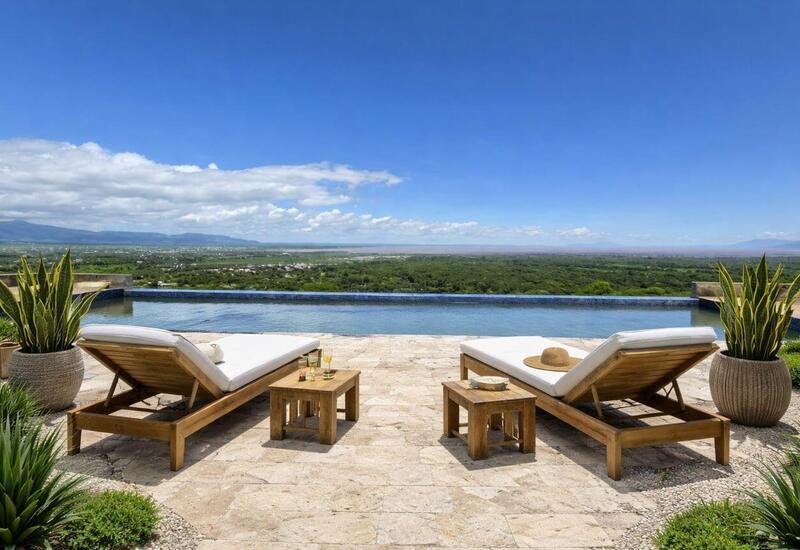 Infinity pool terrace with two wooden sun loungers and side tables, drinks and hat, overlooking green valley and distant mountains under a blue sky