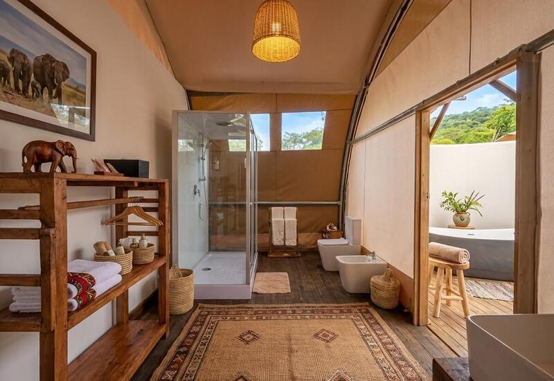 Safari-style glamping bathroom with wood shelving, glass shower, bidet, outdoor freestanding tub on private deck and nature view